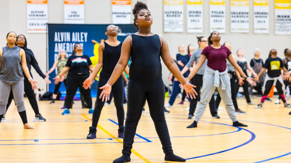 a diverse group of people, mostly women but all ages, participate in a choreography workshop. They stand in a strong pose, feed apart and arms down