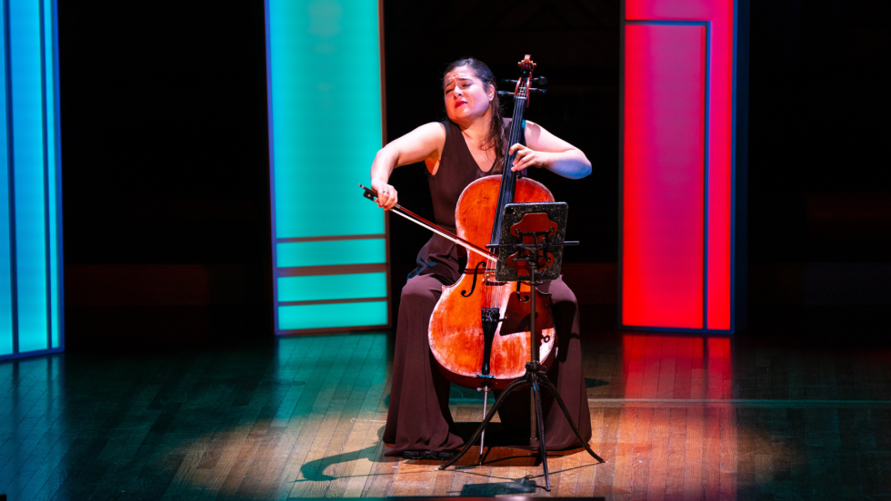 a woman with light skin and long brown hair passionately plays a cello in front of multicolored lighted panels. 