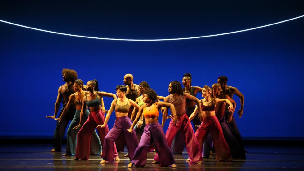 a diverse company of dancers in colorful outfits in a triangle formation lock their right arms out to the sides and arc their left arms behind them