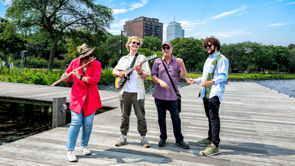 a flutist, a guitarist, an upright bass player and a drummer stand on a dock on the bank of the Charles River in Boston