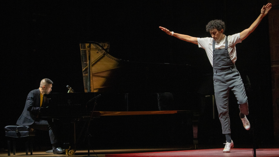 an asian american man plays the piano while a person with dark curly hair and striped coveralls tap dances