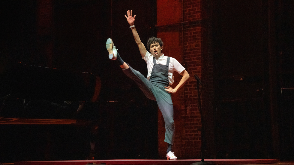 a person with dark curly hair and striped overalls leaps into the air, one arm and one leg extended almost vertical