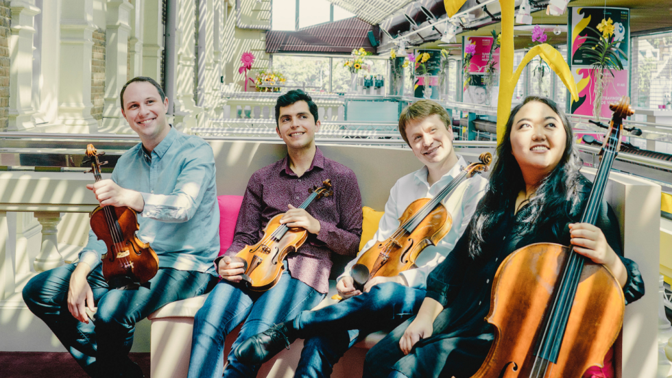 Three men and one woman hold stringed instruments in an atrium decorated with colorful artwork