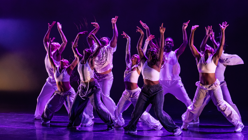 under purple light, a company of Black dancers lunge forward, with one arm bent and raised 