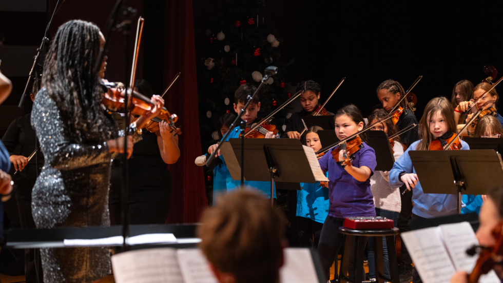 from behind, a black woman in a sparkly silver sheath dress leads a diverse group of elementary and middle school students in a strings workshop.