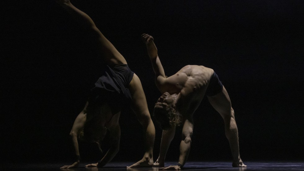 Circa performers in black against black background. One performer does a handstand with their leg in the air while the other is in a left side bend.