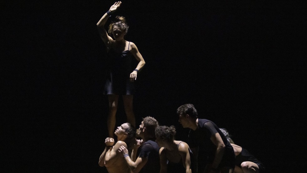 A group of Circa performers in black against a dark black background. Some performers are low to the ground while one dancer stands with their arms in a geometric position.