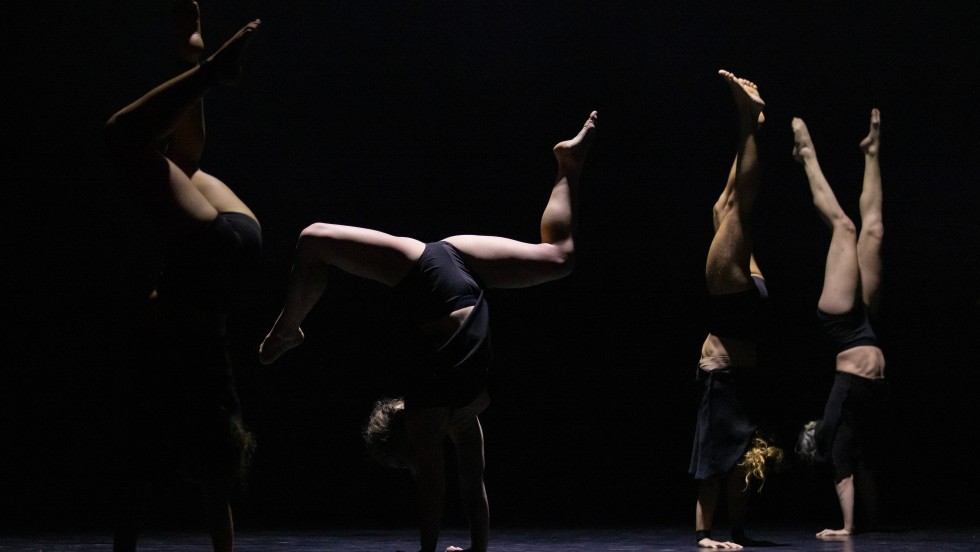 Three Circa performers in black against black background. Two performers do straight handstands on the right while a third does a handstand split on the left. 