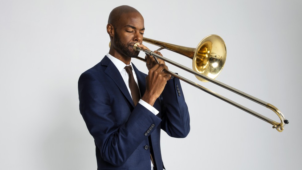 Chris Crenshaw,a black man in a blue suit playing a trombone against a white background. 