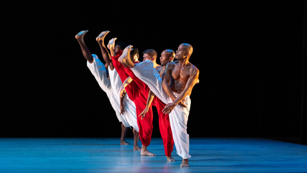 six dancers, the men shirtless in loose white pants and the women in flowing red jumpsuits, in a line, extending upstage. they each extend a straight leg, their feet as high as their heads