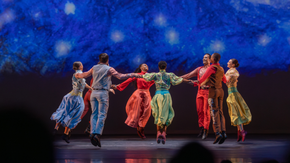 dancers dressed in brightly colored prairie-style dresses clasp arms and dance in a circle