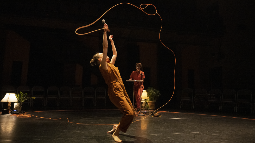 the dancer twirls the cord of a device above her head like a lasso with the musician in the background