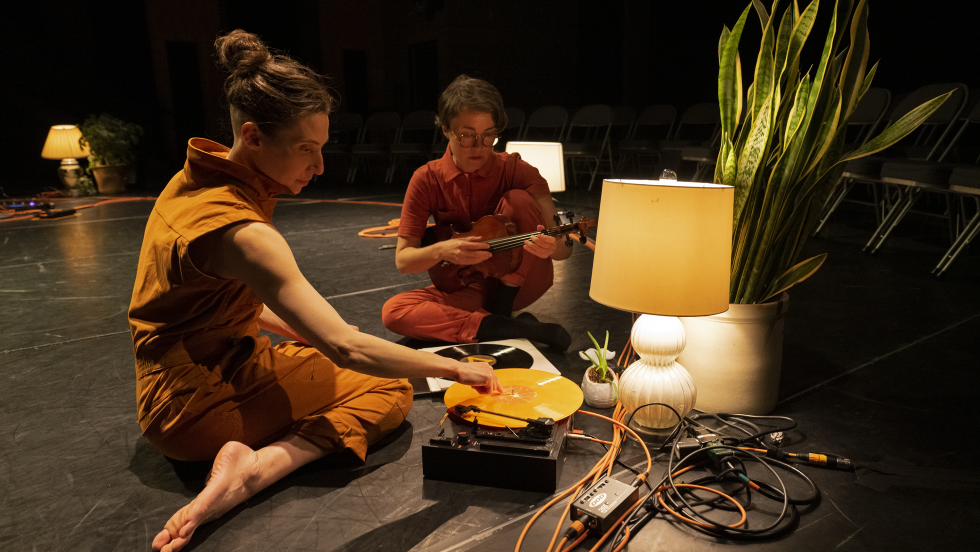 the dancer and the musician crouch and sit on the floor near vintage electroinics, a shaded table lamp, and a tall green plant