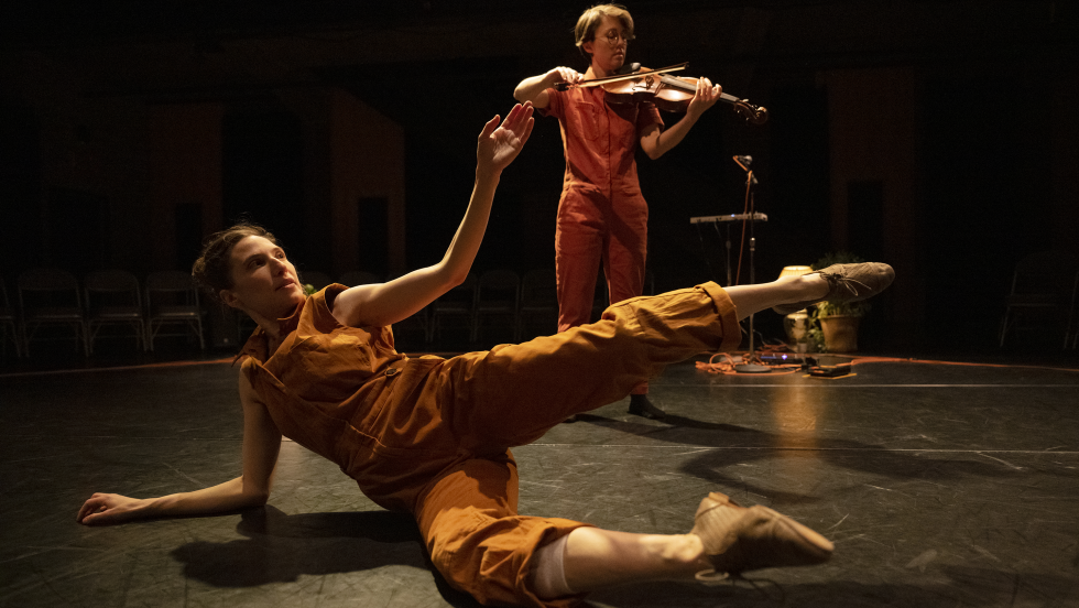 a dancer in an ochre jumpsuit stretches on her side on the floor, while a musician plays a viola behind her