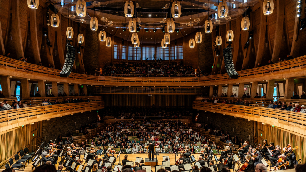 a photo of an oval-shaped concert hall, with light wood structural beams. An orchestra performs on stage.
