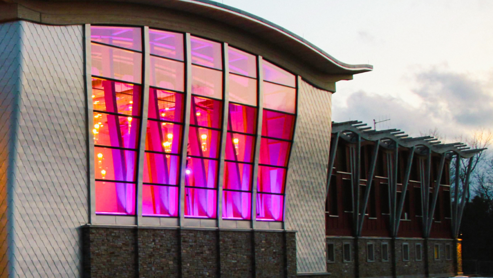 a contemporary performing arts center with an undulating roof. A function room with generous windows is brightly illuminated in red, pink, purple lighting