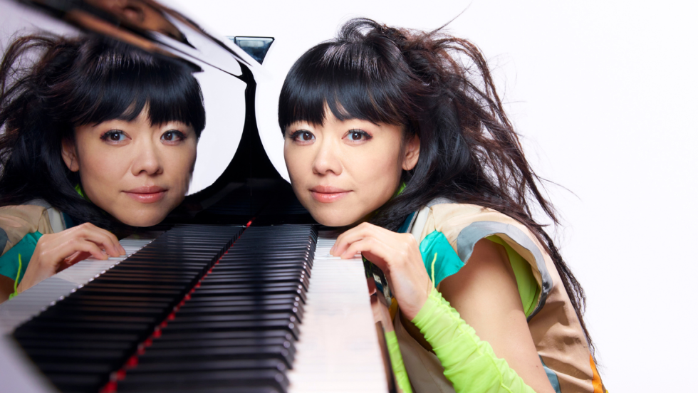 hiromi rests her chin on the keys of a grand piano. She wears a colorful top and green arm warmers