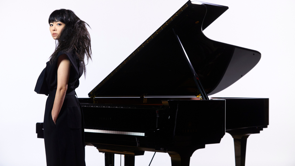 hiromi, a japanese woman wearing black with her hair in a textured ponytaill, stands with her back to a grand piano