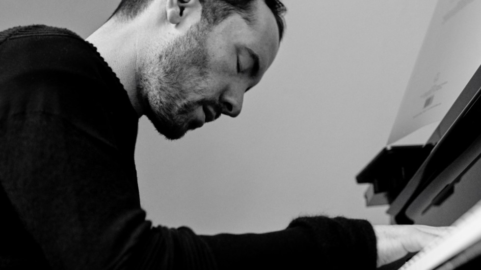 A white man plays the piano wearing a black top in front of a gray background. 