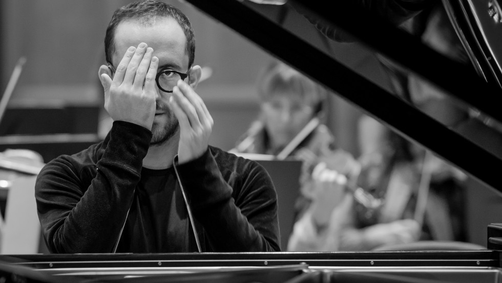 Igor levit, a white man with brown hair wears black glasses and covers one eye with his hand as he sits in front of a piano. 