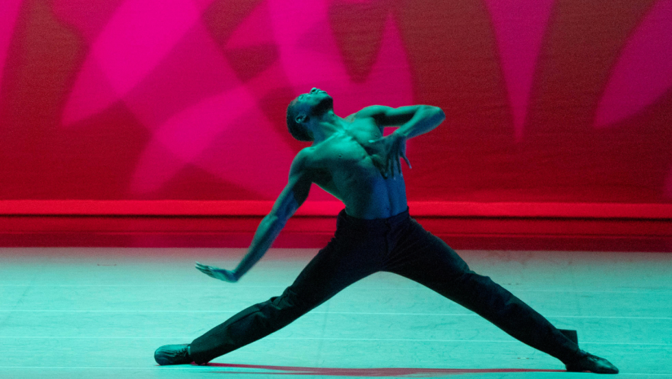 legs making a wide triangle, a dancer, shirtless, his brown skin bathed in an unearthly blue-green light. His face is raised, his torso bends back, one arm extends below with a hand parallel to the floor and his other hand clutching his chest.