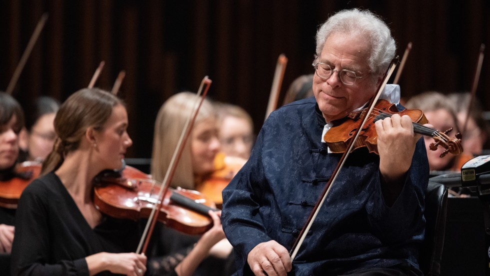 Violinist Itzhak Perlman a man plays a violin with an orchestra behind him. 