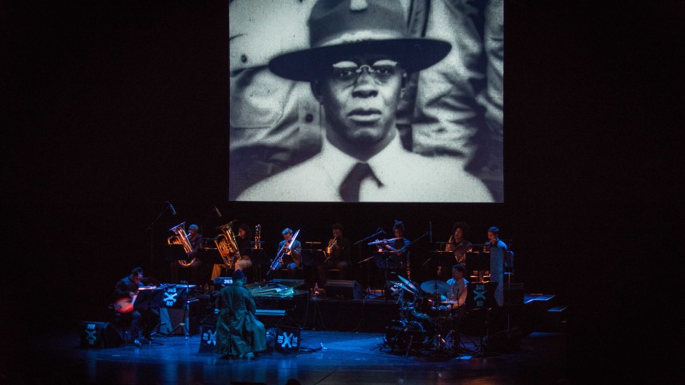 A screen above an ensemble onstage has a black police officer projected on it, while the ensemble plays music below. 