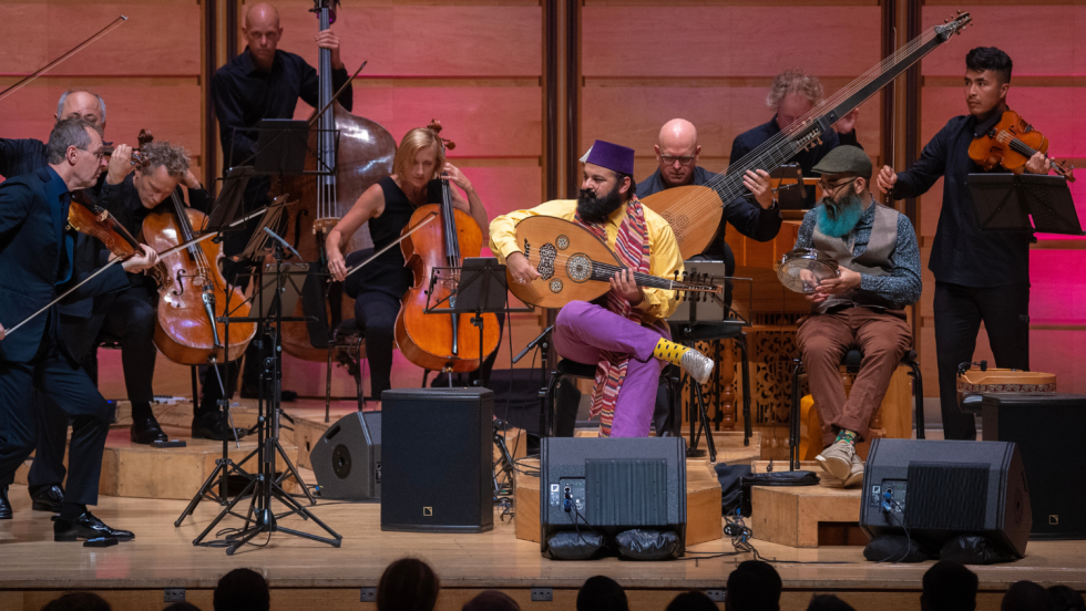 two egyptian australian men perform as guest artists in front of a chamber orchestra. One of the men plays a lute-like oud, the other a round percussion instrument.