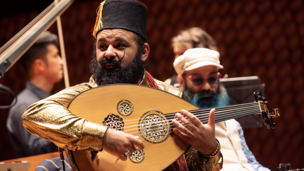 an Egyptian Australian man, with a yellow shirt, a curly mustache, and a long full beard, focuses to his right as he performs on his lute-like oud.