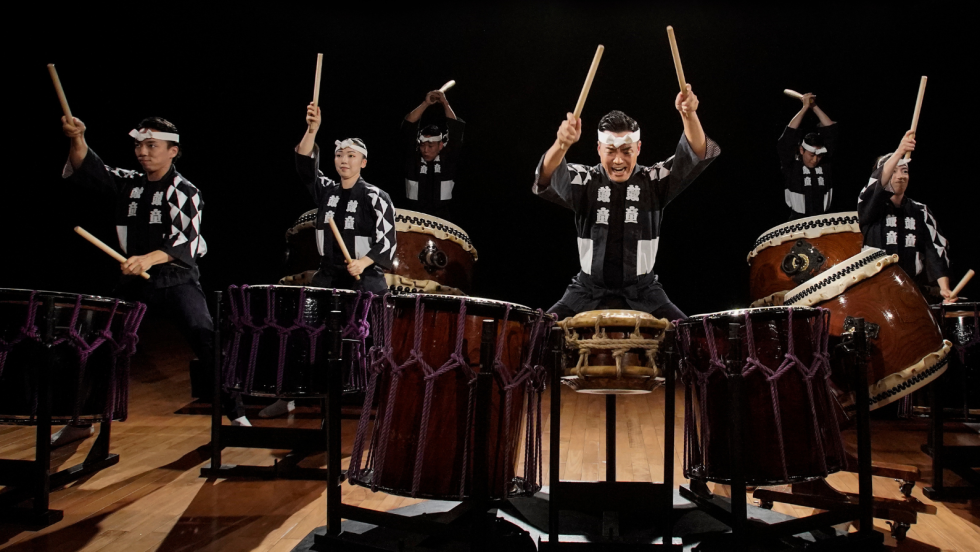 a company of drummers perform together, smiling with their drumsticks raised