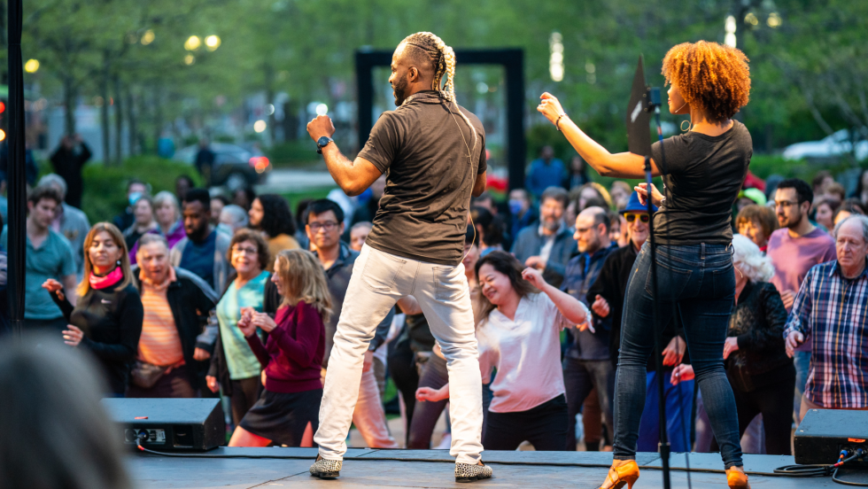 facing away, a man and a woman lead an outdoor dance lesson for a large crowd