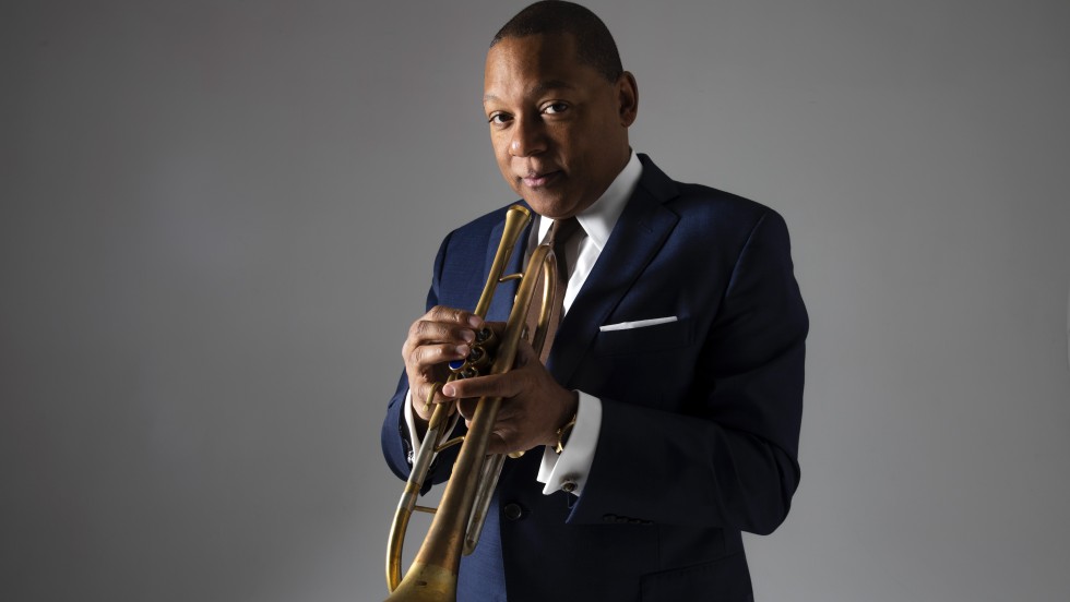 Wynton Marsalis, a black man in his sixties holding a trumpet in a black suit against a gray background.