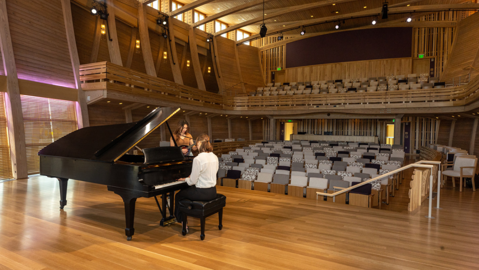 from the stage, a woman plays a grand piano while another woman plays a mandolin in an empty theatre paneled in light wood.
