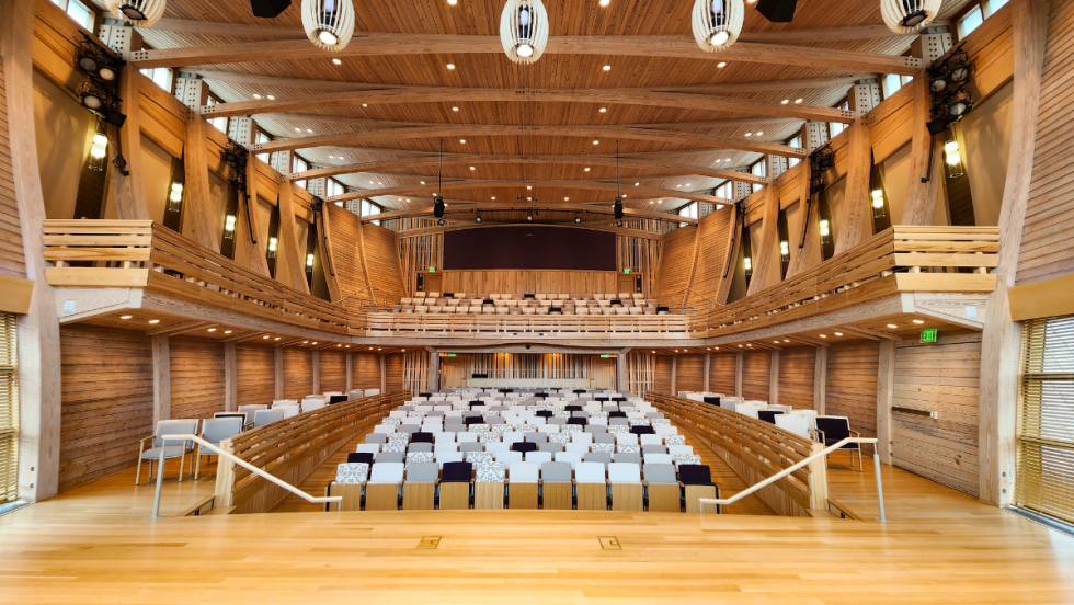 from center stage, a concert hall, with curved walls clad in pine wood. Behind us is a full-height window and the room is airy, bright, and neutral.