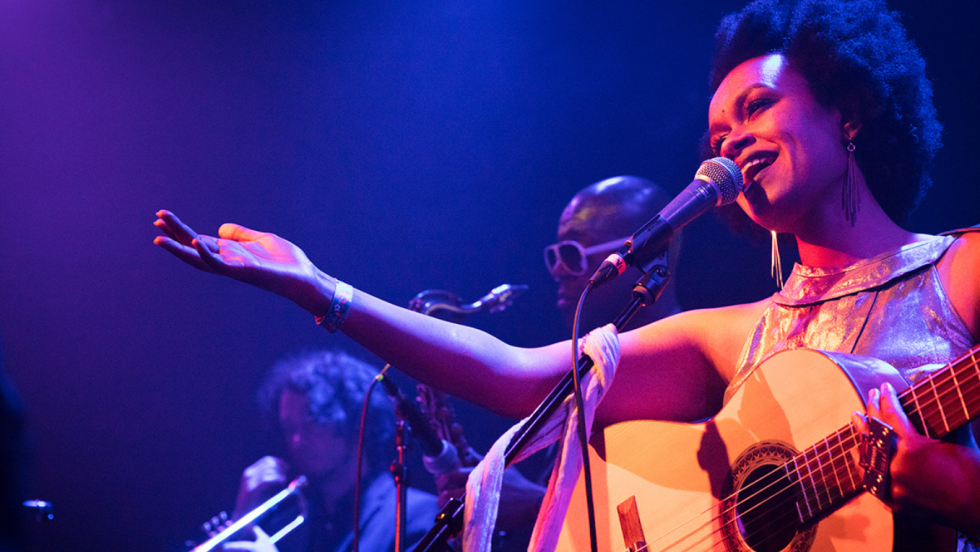 on a stage washed in purple light, meklit extends an arm toward her audience as she sings and plays guitar