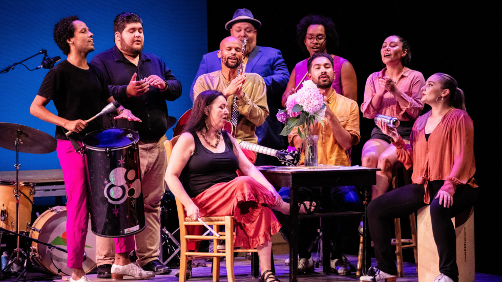 dancers and musicians crowd around an old-fashioned tube TV on a stage set like a house