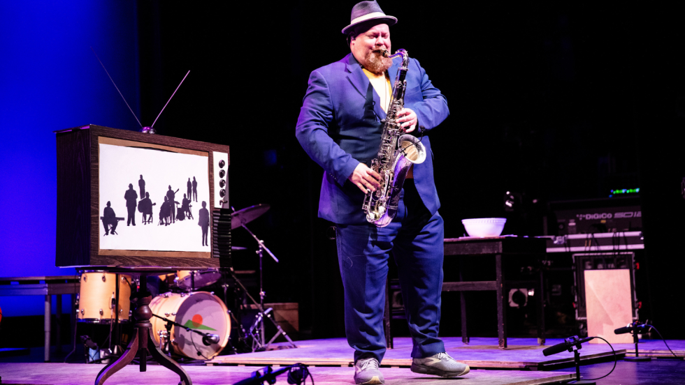 a saxophone player performs alone in a room of a stage set, with a black and white TV in the background