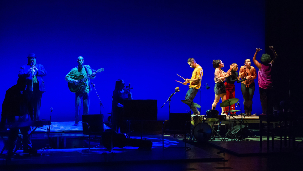 on a stage set with multilevel platforms, a guitarist performs slightly out of the spotlight. Dancers, crowded onto one platform descend, one drumming with sticks as he goes.