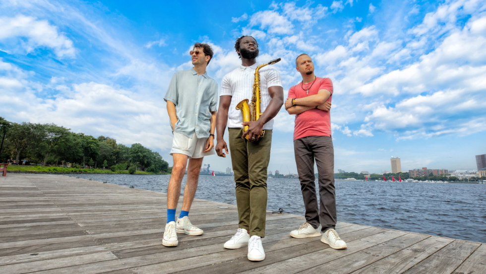 three members of a jazz band, the central person holding his tenor saxophone, stand on a dock in the Charles River. The river and the skyline of Cambridge are behind them.
