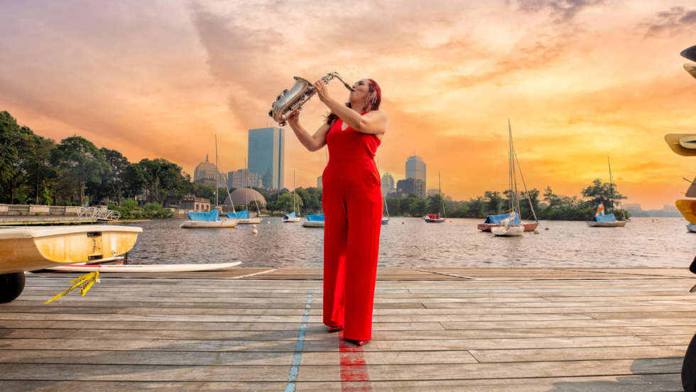 in front of a vivid sunset, a saxophone player performs on a dock in the Charles River. Small boats are on the dock near her, and she wears a vermillion halter-neck jumpsuit.
