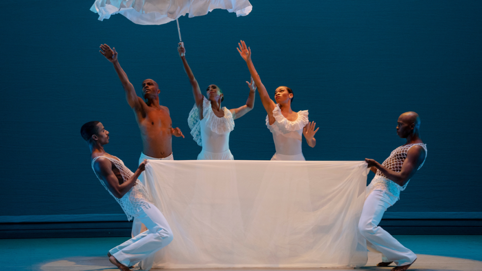 two dancers hold a white fabric in front of three others. the woman in the center raises a white umbrella high