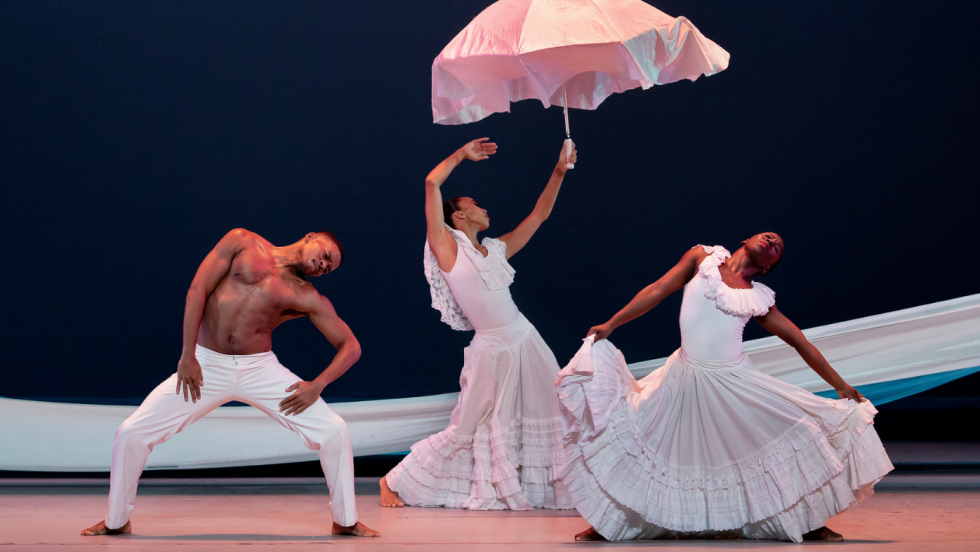 In white garments and underneath a fluttering white umbrella, three dancers lean and gesture to their left, knees bent, in a strong but flexible posture.