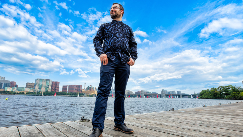 a low-angle photo of a dark-haired man with a short beard on a dock in the Charles River. He wears a dark-patterned shirt and stands with his thumbs hooked in his pockets. the river and Cambridge are behind him.
