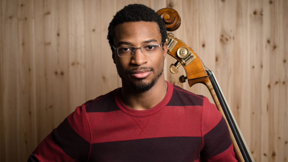 a young black man with a striped sweater and a closed smile holds the neck of his double bass