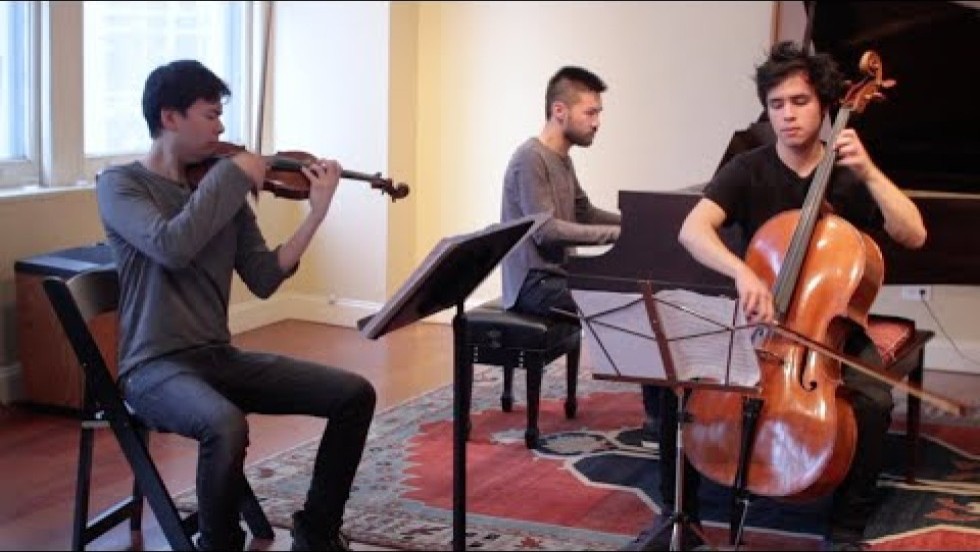 three young men play Charles Ives' Piano Trio, Movement 2 in a sunny room that overlooks a city street. They play a violin, a cello, and a piano in a brightly lit 