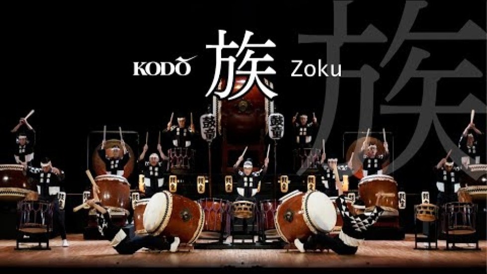 A group of ten male and female drummers in black and white long-sleeve outfits play KODŌ's “Zoku” on various types of Japanese drums