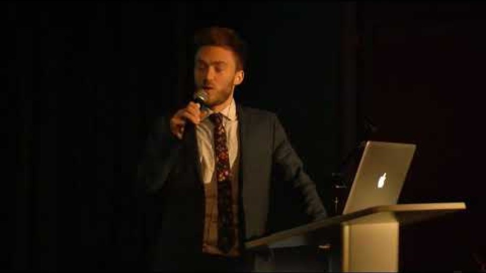 a young man at a lectern with an open laptop gives a presentation with a slideshow featuring photos and music notation