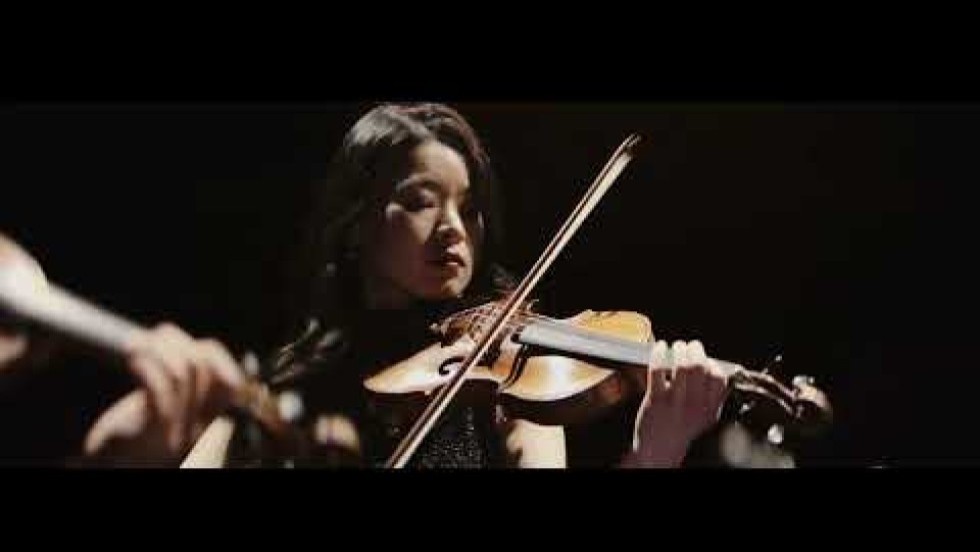 A string quartet, all in black, dramatically spotlit on a dark stage. The camera circles around them as they perform vigorously.