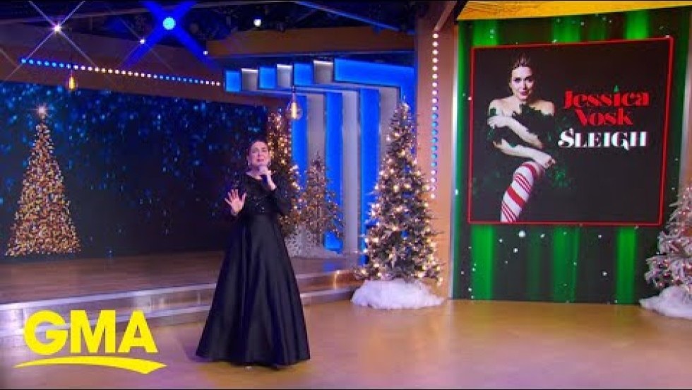 a white woman in a floor length black dress performs on a stage flanked by christmas trees and other holiday decor