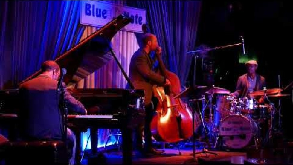 a piano trio, on a blue-draped stage with a sign reading Blue Note. we are behind the pianist 